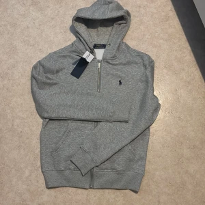 Grå hoodie från Polo Ralph Lauren - Säljer en klassisk helt ny grå hoodie från Polo Ralph Lauren med dragkedja och huva. Tröjan har ribbade muddar, fickor framtill och den ikoniska broderade loggan på bröstet. Perfekt för en avslappnad stil. Snören till tröjan finns. Pris kan diskuteras vid snabb affär 