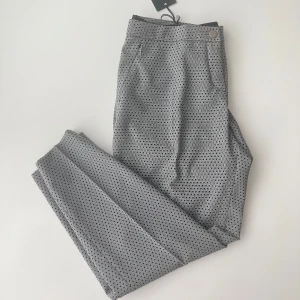 Grå mönstrade kostymbyxor från Rosner - Snygga grå kostymbyxor från Rosner med diskret geometriskt mönster. Byxorna har slim passform, midwaist och klassiska fickor fram samt passpoalfickor bak.