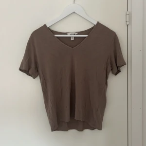 Brun v-ringad t-shirt från H&M - Säljer en enkel och stilren beige t-shirt från H&M med v-ringning. T-shirten har korta ärmar och är tillverkad i mjuk bomull, perfekt för en avslappnad look.