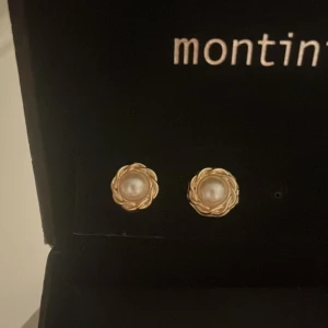 Guldiga örhängen med pärldetalj  - Säljer ett par eleganta örhängen från Montini med guldtonad ram i blomform och vit pärla i mitten. Perfekta för att ge en klassisk touch till din stil.