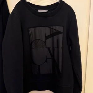 Svart sweatshirt från Calvin Klein Jeans - Säljer en svart sweatshirt från Calvin Klein Jeans med stor, glansig logga framtill. Tröjan har rund halsringning och långa ärmar. Perfekt för en stilren och avslappnad look.