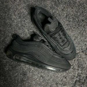Nike Air Max 97 Triple Black - Säljer ett par helsvarta Nike Air Max 97 med ikonisk vågig design och synlig luftbubbla i sulan. Skorna har snörning och är tillverkade i en mix av syntet och mesh för bra komfort och stilren look.