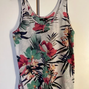 Blommig linnetopp från H&M - Säljer en blommig linnetopp från H&M i vitt med stora färgglada blommor i rött, grönt och gult. Toppen har ärmlös design och rundad halsringning. Perfekt för varma dagar och enkel att matcha med jeans eller shorts.