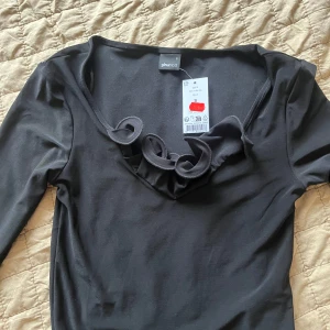 Svart topp med volangdetalj från Gina Tricot - Säljer en svart långärmad topp från Gina Tricot med snygg volangdetalj längs den v-ringade halsen. Toppen är i ett mjukt material och passar perfekt till många olika outfits.helt oanvänd 