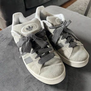 Grå Adidas Campus sneakers i mocka - Säljer ett par grå Adidas Campus sneakers i mocka med klassiska tre svarta ränder på sidan och vit sula. Skorna har grå snörning och Adidas-logga på plösen. Perfekta för en stilren vardagslook.
