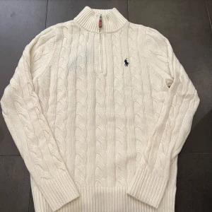 Vit stickad tröja med dragkedja från Polo Ralph Lauren - Kabelstickad vit tröja från Polo Ralph Lauren med halv dragkedja och broderad logga på bröstet. Tröjan har lång ärm, hög krage och ribbade muddar. Perfekt för kyligare dagar.
