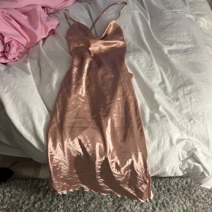 Champagnefärgad satinklänning med smala axelband - Säljer en glansig champagnefärgad långklänning i satin med smala axelband och snygga knytband i ryggen. Klänningen har en elegant och enkel design som ger en lyxig känsla. Perfekt för speciella tillfällen!