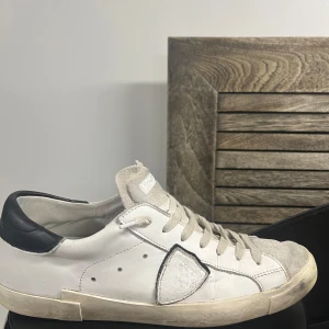 Vita sneakers från Philippe Model - Säljer ett par vita Philippe Model sneakers med svarta detaljer vid hälen och klassisk snörning. Skorna har en rund tå och platt sula, samt diskret logotyp på sidan. Perfekta för en stilren och avslappnad look.