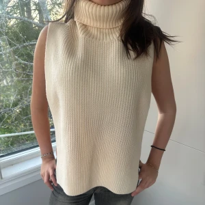Beige stickad väst med polokrage - Super snygg beige stickad väst med hög, ribbad polokrage. Västen är ärmlös och öppen i sidorna. Aldrig använd men saknar prislapp.