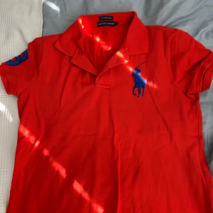 Röd pikétröja från Ralph Lauren - Röd pikétröja från Ralph Lauren med blå logga broderad på bröstet och siffra på ärmen. Klassisk krage och korta ärmar, modellen är 'The Skinny Polo'. Perfekt för en sportig och stilren look. Pris kan diskuteras 