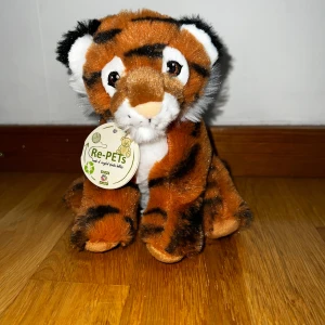 Mjukisdjur tiger från Re-PETS - Säljer ett gulligt mjukisdjur i form av en tiger från Re-PETS. Tigern är orange med svarta ränder, vita detaljer på ansikte och öron samt har en lång svans. Pälsen är mjuk och fluffig, perfekt som gosedjur eller dekoration. Helt ny och oanvänd. Har fortfarande lappen kvar 