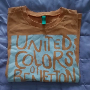 Orange t-shirt från United Colors of Benetton - Snygg barn orange t-shirt med stort blått tryck framtill där det står 'United Colors of Benetton'. Klassisk rund hals och korta ärmar. Perfekt för en avslappnad stil.