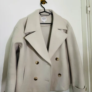 Beige kappa från Kappahl - PASSAR M-L- XL Stilren beige kappa från Kappahl med bred krage och dubbelknäppning med fyra knappar framtill. Jackan har lång ärm och en klassisk, rak passform. Perfekt för dig som gillar en tidlös look.
