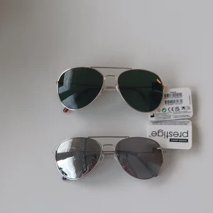 Aviator solglasögon från Foster Grant - Två par klassiska aviator-solglasögon från Foster Grant. Ett par med gröna glas och guldfärgad metallbåge, och ett par med gråtonade glas och silverfärgad metallbåge. Båda har tunna skalmar och näsbrygga i metall.