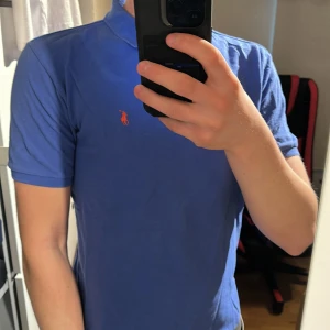 Blå pikétröja från Ralph Lauren - Säljer en klassisk blå pikétröja från Ralph Lauren med orange broderad logga på bröstet. Tröjan har korta ärmar, krage och två knappar vid halsen. Perfekt för en stilren och avslappnad look. M men känns som S. Äldre taggen 