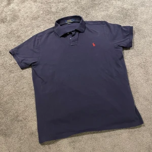 Mörkblå pikétröja från Polo Ralph Lauren - Klassisk mörkblå pikétröja från Polo Ralph Lauren med röd broderad logga på bröstet. Tröjan har krage och korta ärmar, tillverkad i bomull för en bekväm känsla. Perfekt för en stilren och avslappnad look.