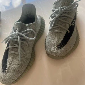 Adidas Yeezy Boost 350 V2 i ljusgrå - Säljer ett par Adidas Yeezy Boost 350 V2 i ljusgrå med svart detalj på sidan och text. Skorna har stickad ovandel, snörning och en tjock, räfflad sula i gummi. Perfekta för dig som gillar streetwear och vill ha något unikt.