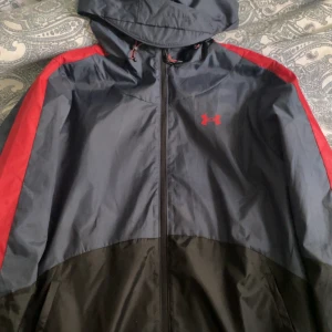 Under Armour jacka  - Säljer en snygg vindjacka från Under Armour i svart och grått med röda detaljer och logga. Jackan har huva, dragkedja och ribbade muddar. Perfekt för träning eller utomhusaktiviteter.