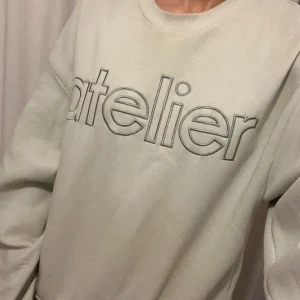 Vit sweatshirt med 'atelier'-tryck - Säljer en vit sweatshirt med stort broderat 'atelier'-tryck på bröstet. Tröjan har rund halsringning och långa ärmar. Perfekt för en avslappnad och stilren look.