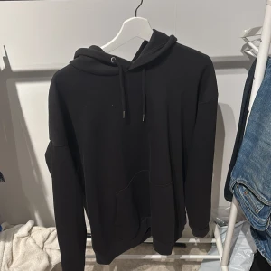 Svart hoodie med ficka - Säljer en klassisk svart hoodie med huva och dragsko. Tröjan har en stor magficka och långa ärmar. Perfekt för en avslappnad stil och enkel att matcha med jeans eller joggers.