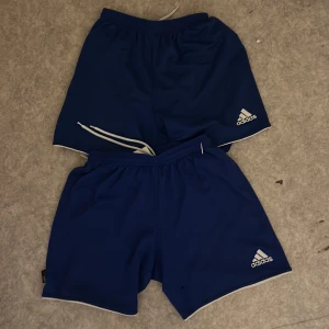 Blå träningsshorts från Adidas - Två par blå träningsshorts från Adidas med vit logga på vänster ben och elastisk midja. Klassisk sportig look med enkel design, perfekta för  fotbollen eller annan sport / för barn, 120 för båda