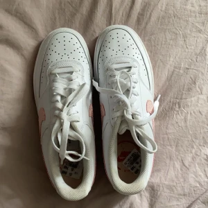 Nike Air Force 1 med rosa detaljer - Säljer ett par vita Nike Air Force 1 med ljusrosa detaljer, helt nya med prislapp kvar