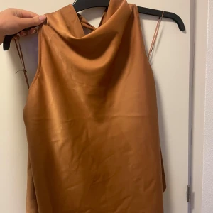 Satintopp med knytband  - Säljer en snygg brun satintopp med halterneck och långa knytband i ryggen. Toppen har en draperad halsringning. Perfekt för till fest eller middagar. Använd 2-3 gånger. Färgen är svår att beskriva men något åt det brun/orange/gula