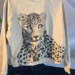 Beige sweatshirt med leopardtryck från H&M - Cool beige sweatshirt från H&M med stort leopardmotiv framtill och texten 'ALWAYS RUN WILD' på ena ärmen. Tröjan har rund hals och långa ärmar, perfekt för en avslappnad stil. Har lite svarta färgfläckar på