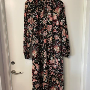 Blommig långklänning från H&M - Säljer en svart långklänning från H&M med färgglatt blommönster i rosa, beige och grönt. Klänningen har lång ärm och hög hals med knytning. Perfekt för dig som gillar bohemisk stil och vill sticka ut.