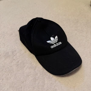 Svart keps från Adidas med broderad logga - Svart keps från Adidas med vit broderad Trefoil-logga framtill och baktill. Justerbar rem baktill för perfekt passform. Klassisk och stilren design som passar till det mesta.