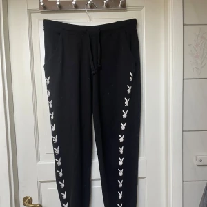 Svarta mjukisbyxor med Playboy-logga - Svarta mjukisbyxor med vita Playboy-kaniner längs båda benen. Byxorna har dragsko i midjan och sidofickor. Perfekta för en avslappnad och trendig look.