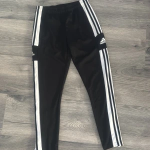 Svarta träningsbyxor från Adidas med vita ränder - Svarta träningsbyxor från Adidas med klassiska vita ränder längs sidorna och Adidas-logga på låret. Byxorna har elastisk midja och är tillverkade i ett syntetmaterial som passar perfekt för träning.