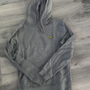 Grå hoodie från Lyle & Scott - Säljer en klassisk grå hoodie från Lyle & Scott med gul logga på bröstet. Tröjan har huva med dragsko och en stor magficka framtill. Perfekt för en avslappnad stil.