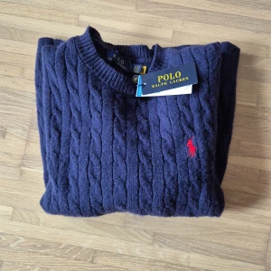 Stickad ralph lauren + tyg armband i min profil  - Mörkblå kabelstickad tröja från Polo Ralph Lauren med rund halsringning och röd broderad logga på bröstet. Klassisk modell med ribbade muddar vid ärmslut och nederkant. Köp nu och få med ett tyg armband i min profil!