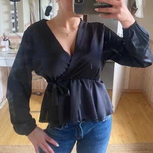 Svart omlottblus med knytband - Svart omlottblus med v-ringning och knytband i midjan. Långa ärmar med lätt puff och resår vid ärmslut. Snygg och stilren modell som passar till jeans eller kjol.