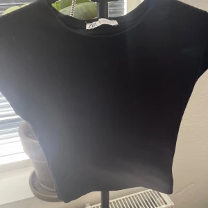 Svart croppad topp från Zara - Säljer en svart croppad topp från Zara. Enkel och stilren design med rund halsringning och korta ärmar. Perfekt att matcha med både jeans och kjol.
