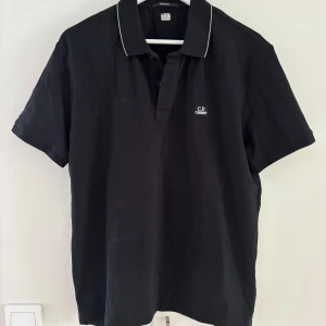 Svart pikétröja från C.P. Company - Svart pikétröja från C.P. Company med vit piping på kragen och liten logga broderad på bröstet. Klassisk polokrage med knappar framtill. Perfekt för en clean och stilren look.
