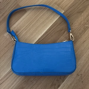 Blå axelväska med krokodilpräglat mönster - Säljer en trendig blå axelväska med krokodilpräglat mönster och guldfärgade metalldetaljer. Väskan har en enkel, stilren design och ett avtagbart axelband. Perfekt för att addera färg till din outfit!