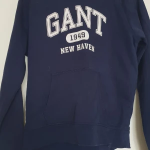 Mörkblå hoodie från Gant - Säljer en mörkblå hoodie från Gant med vit text 'GANT 1949 NEW HAVEN' på bröstet. Tröjan har huva, magficka och långa ärmar. Perfekt för en avslappnad stil.
