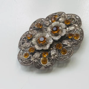 Vintage brosch i silverfärg med orange stenar - Unik brosch i silverfärgad metall med detaljerade blommor och gnistrande orange stenar. Två saknas.  Vacker filigran design med vackra löv och blomdetaljer. Perfekt som accessoar för att lyfta din outfit.