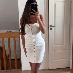 Vit knappklänning i jeansmaterial - Säljer en vit, tight klänning i jeansmaterial med knappar framtill. Klänningen är ärmlös och har en figurnära passform som framhäver midjan. Perfekt för sommarens alla tillfällen!