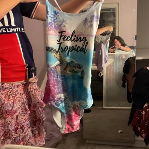 Baddräkt med tropiskt tryck och text - Säljer en färgglad baddräkt med motiv av havssköldpadda, palmer och texten 'Feeling Tropical' framtill. Baddräkten har smala axelband som korsas i ryggen och en blå, rosa och vit färgskala. Perfekt för strand eller pool!