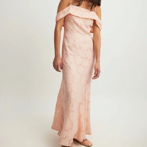 Ljusrosa axelbandslös långklänning - Elegant ljusrosa långklänning med offshoulder och smala axelband. Klänningen har ett subtilt blommigt mönster och en lätt utsvängd passform. Perfekt för bal eller festliga tillfällen.