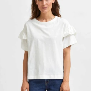 Vit t-shirt med volangärmar - Säljer en vit t-shirt med rund hals och volangdetaljer på ärmarna. T-shirten har en rak passform och är tillverkad i mjuk bomull. Som ny