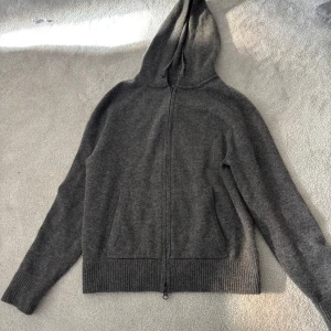 Grå kashmir hoodie med dragkedja - Mysig grå hoodie i 100% kashmir från Singular Society. Tröjan har dragkedja framtill, huva med snören och ribbade muddar vid ärmslut och nederkant. Perfekt för lager-på-lager och en avslappnad stil.