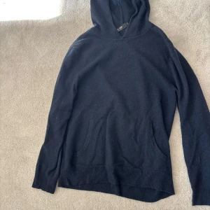 Svart hoodie i ull från LISA YANG - Mysig svart hoodie från LISA YANG i 100% cashmere. Tröjan har huva, lång ärm och en stor magficka framtill. Perfekt för kyliga dagar och enkel att matcha med det mesta.