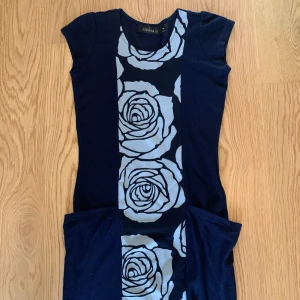 Mörkblå klänning med stora blommor från Mina UK - Supersnygg svart klänning från Mina UK med stora vitblommiga mönster framtill. Klänningen har korta ärmar och två stora fickor på sidorna. Perfekt för dig som gillar unika detaljer och vill sticka ut.