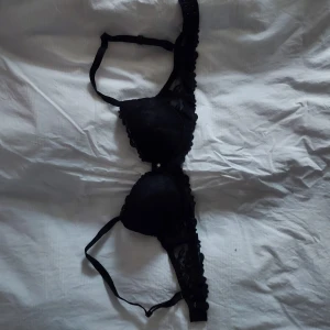 Svart spets-bh - Elegant svart bh med spetsdetaljer och vadderade kupor. Den har justerbara axelband och en liten dekorativ pärla framtill. Perfekt för en stilren look.