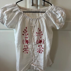 Vit blus med broderade röda blommor och knytning - Säljer en vit blus med puffärmar och vacker röd brodyr framtill. Blusen har knytning längs framsidan och en luftig, romantisk känsla. Perfekt för dig som gillar boho-stil och detaljerade mönster.