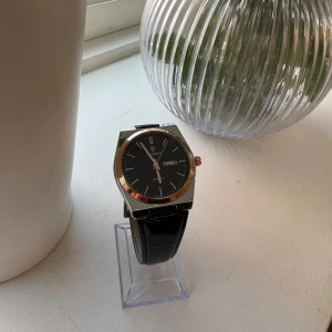 Svart herrklocka från Poedagar - Stilren herrklocka från Poedagar med svart urtavla, silverfärgat och roséguldfärgat boett samt svart läderarmband med krokodilstruktur. Klockan har dag- och datumvisning och klassisk design.